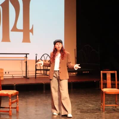 Girl in flat cap standing on stage in front of 1914 screen